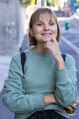 Smiling young woman standing on city sidewalk wearing green sweatshirt and backpack holding coffee