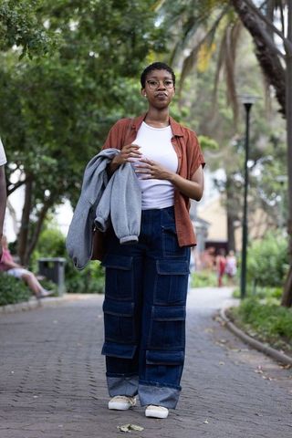 African woman walking in park holding gray sweatshirt while wearing glasses and cargo jeans