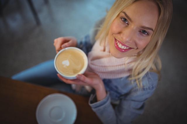 Blonde woman smiling while holding a cup of coffee with latte art. Ideal for lifestyle blogs, coffee shop promotions, social media content, and advertising related to cafés and beverages.