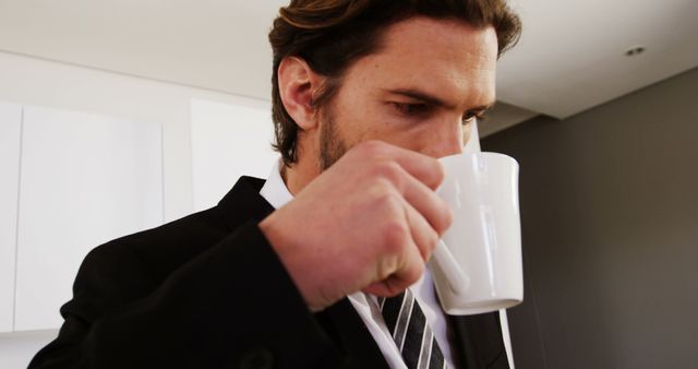 Businessman in suit drinking coffee during workday inside modern office. Perfect for corporate environment themes, morning routine materials, refreshment concepts, and professional lifestyle promotions.