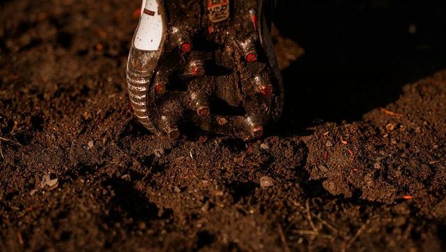 Close-up muddy soccer cleat pressing into soil with red-tipped studs catching warm light