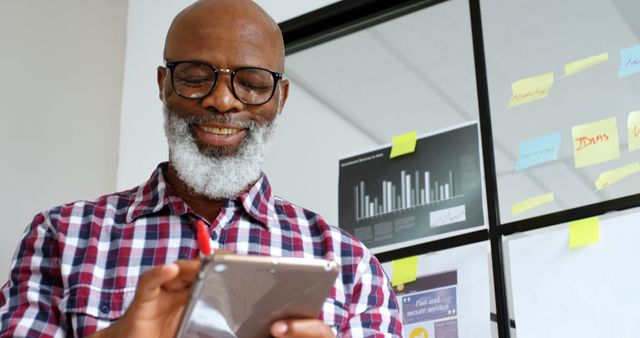 Senior man with gray beard and glasses smiling while using a tablet in an office with charts and sticky notes on the wall in the background. Ideal for business, technology, productivity, and modern workspace concepts.