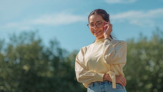 Smiling young woman adjusting glasses in sunny park wearing pullover and high-rise jeans