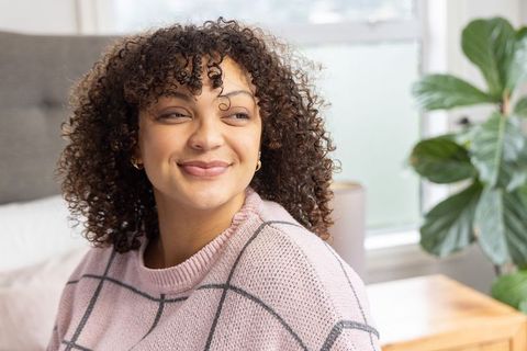 African american woman smiling and looking right in cozy bedroom wearing pink sweater