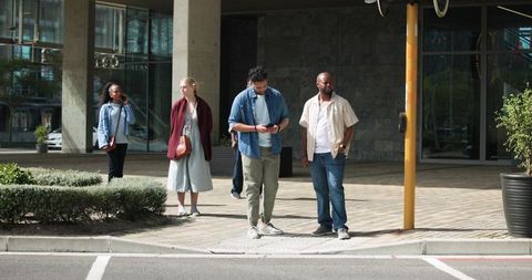 Multicultural pedestrians waiting at curb and checking smartphones near modern office plaza