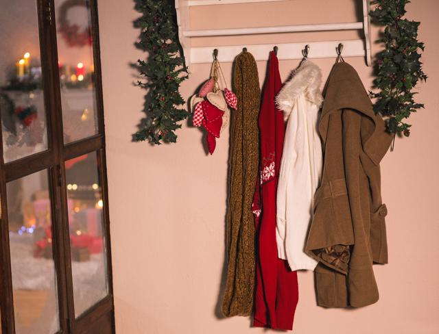 Winter coats and scarves hanging on wall hooks in a cozy home interior. The scene is decorated with festive holiday elements, creating a warm and inviting atmosphere. Ideal for use in articles or advertisements about winter fashion, home decor, holiday preparations, and seasonal lifestyle.