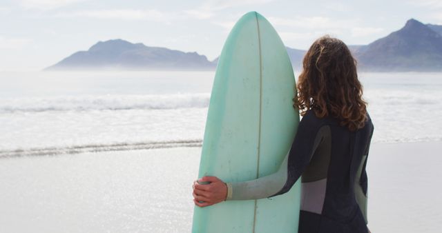 Woman wearing wetsuit embracing surfboard while standing on sandy beach, ocean waves gently rolling in, mountains in background. Perfect for topics related to outdoor sports, surfing, beach lifestyle, female empowerment, vacation destinations.