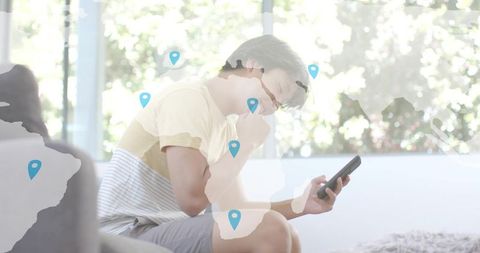 Young asian man sitting and using smartphone in bright home with global map pins overlay