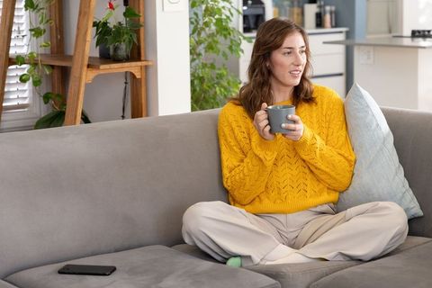 Woman sipping coffee on sofa wearing yellow sweater relaxing at home with smartphone nearby
