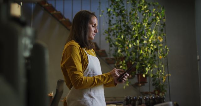 Female barista in a modern cafe using a tablet. She is wearing a yellow top and a white apron, surrounded by green plants. Ideal for content related to small businesses, modern cafes, technology integration in workplaces, and the coffee shop industry.