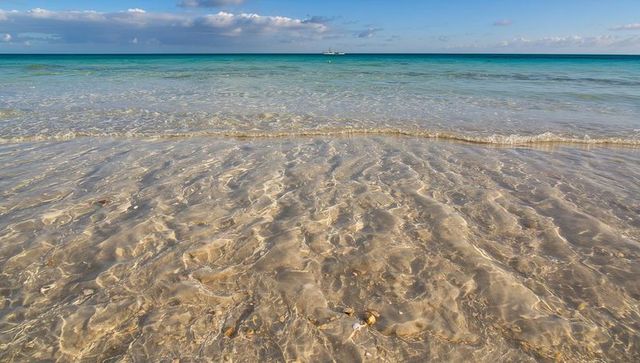 Shimmering clear shallow seawater over rippled sand at turquoise shoreline with distant boat