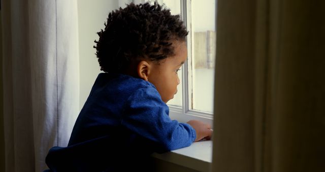Side view of cute little black boy leaning on window sill in a comfortable home. He is looking outside through window 4k