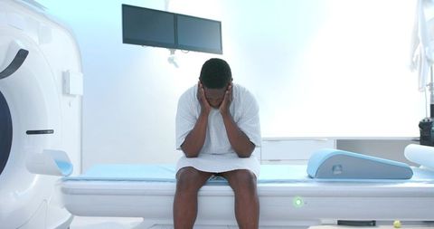 African american man sitting on ct scanner table covering face experiencing anxiety