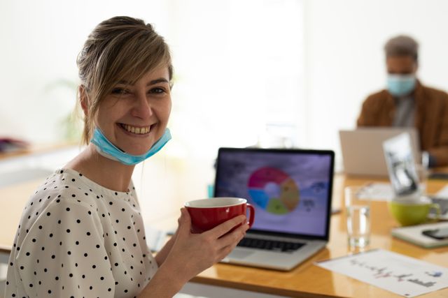 Caucasian woman with her facemask pulled down smiling while holding a cup of coffee at a meeting in an office. beside her on the desk is a laptop with a graph on the screen.