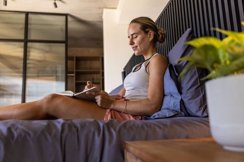 Young woman journaling on bed in modern bedroom with natural light and indoor plant