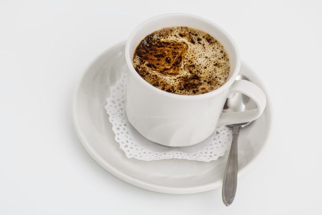 The close-up captures a hot coffee in a white cup and saucer with a spoon, placed on a lace doily against a clean, white background. Perfect for use in articles or advertisements related to coffee shops, breakfast, or beverages. Ideal for websites, blogs, or social media posts that focus on coffee culture, morning routines, or refreshing drinks.