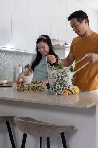 Asian couple unpacking mesh grocery bags at modern kitchen island, sorting fresh produce