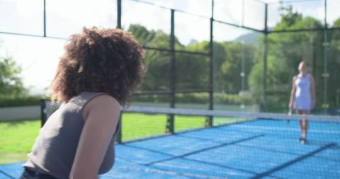 Diverse women leaning and walking to net on blue outdoor court holding rackets, active match