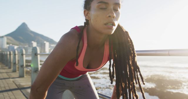 Young woman with long braids taking a break during an outdoor workout by the ocean. This image is ideal for promoting fitness programs, urban lifestyle themes, workout gear, and health and wellness campaigns. Perfect for social media posts, blog articles about fitness journeys, or advertisements for athletic apparel.