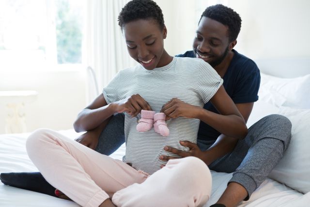 Expecting couple sitting on bed holding baby socks, symbolizing anticipation and joy of upcoming parenthood. Ideal for use in parenting blogs, maternity articles, family planning resources, and advertisements related to baby products or family services.