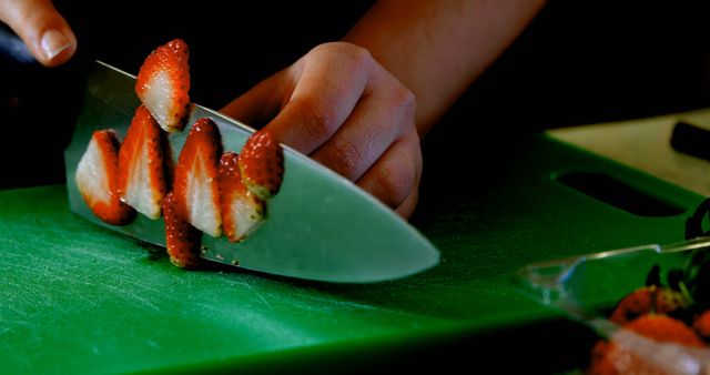 Person slicing fresh strawberries with a knife on a green cutting board. Suitable for food blogs, culinary websites, cooking tutorials, and kitchen product advertisements.