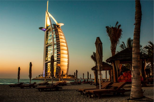 Sunset view of the iconic Burj Al Arab hotel with sun chairs and umbrellas on the beach. The luxurious ambiance and stunning architecture make it an ideal graphic for travel brochures, tourist guides, and marketing materials focused on luxury vacations or Dubai tourism. Perfect for social media posts to highlight beautiful destinations and luxury travel.