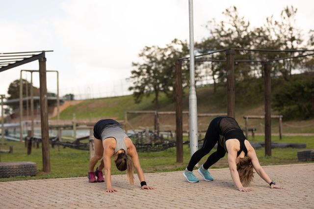 Front view of two Caucasian women wearing sports clothes standing on all fours and stretching at an outdoor gym before a bootcamp training session