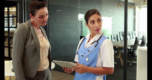 A middle-aged Caucasian nurse is holding a tablet and discussing with a businesswoman in a modern office setting, with copy space. Their expressions suggest a professional exchange of information, related to healthcare services or corporate wellness programs.