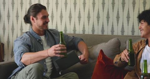 Two friends relaxing on couch sharing beer and laughs with tablet for casual home hangout