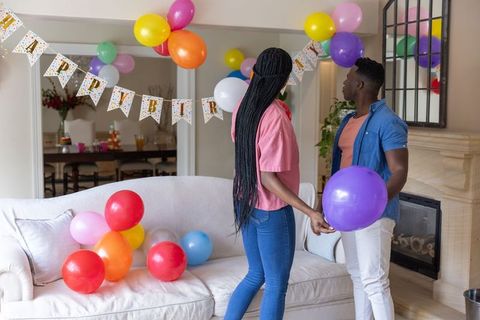 Couple preparing birthday celebration at home holding balloons under birthday banner
