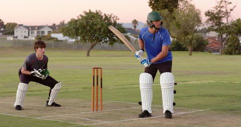 Batsman preparing shot on local cricket pitch at dusk with wicketkeeper ready wearing helmet and pad