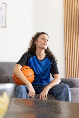 Young woman holding basketball on gray sofa in blue athletic shirt in minimalist living room