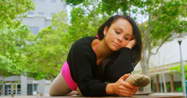 This image captures a dancer stretching outdoors in an urban park. She appears focused and at ease, leaning forward and holding her foot. Suitable for use in promoting fitness routines, dance schools, healthy lifestyle articles, and relaxation techniques.