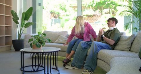 Young couple having relaxed conversation on modern sunlit living room sofa with plants