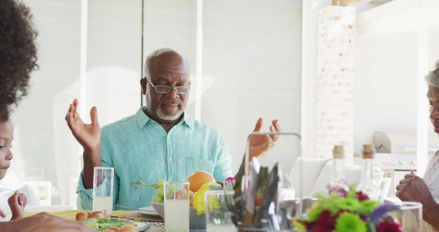 Senior man leading family in prayer around dining table before meal. Ideal for depicting themes of family togetherness, traditional values, gratitude, and intergenerational bonding. Perfect for use in advertisements, religious articles, and community brochures.