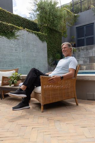 Mature man relaxing on wicker armchair at modern poolside patio with greenery and watch