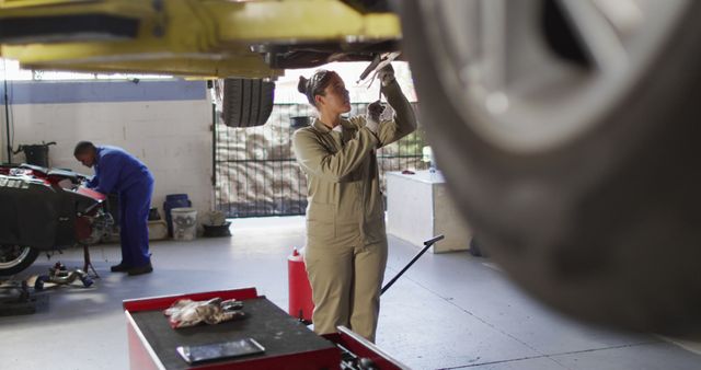 Woman mechanic in uniform working under an elevated car in auto repair shop. Professional setting with tools and equipment visible. Ideal for illustrating automotive services, women in skilled trades, vehicle maintenance activities, and promoting gender diversity in technical professions.