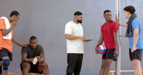 Coach leading diverse male volleyball team during indoor practice with tablet, players chatting