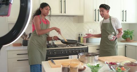 Couple cooking together in modern bright kitchen wearing olive aprons stirring pan