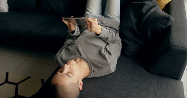 A person is lying upside down on a couch, using a tablet to play a game or watch content. The casual setting and relaxed posture make it suitable for illustrating leisure activities, home technology use, and modern digital lifestyles.
