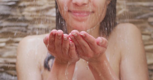 This image shows a woman enjoying a fresh outdoor shower in a garden environment. She has her hands cupped to catch water, which indicates a sense of refreshment and tranquility. This serene and refreshing scene can be used in marketing campaigns for spas, wellness centers, skincare products, or eco-friendly outdoor activities. It is perfect for illustrating themes of purity, health, and relaxation.
