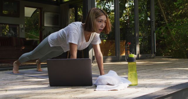 A woman is in a plank position while following an online fitness class on her laptop at home. She is working out indoors near large windows in a well-lit space with a water bottle and towel nearby. Ideal for use in articles or advertisements related to healthy lifestyles, home workouts, online fitness programs, virtual training, and modern fitness trends.