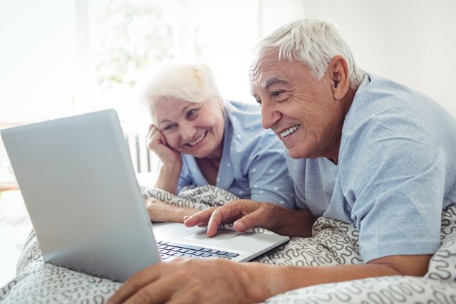 Senior couple lying in bed and using a laptop, smiling and enjoying their time together. This image can be used for promoting technology use among older adults, retirement lifestyle, online communication, and senior living. Ideal for websites, blogs, and advertisements related to senior care, technology for seniors, and family bonding.