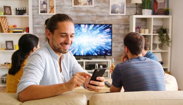Smiling man using smartphone on sofa with friends watching TV casual home streaming moment