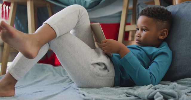 Young boy laying on a comfortable couch with pillows, holding a digital tablet, focusing intently. Perfect for marketing materials related to educational technology, children's leisure activities, screen time balance, or home lifestyle products.