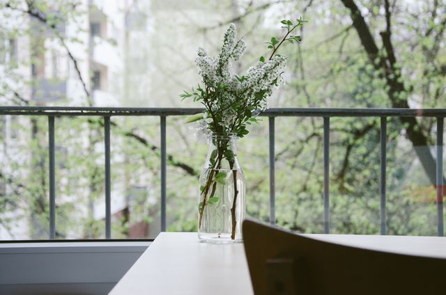 Minimalist vase with green sprigs and white flowers sitting on a table near a window with an outdoor view of greenery and trees. This image can be used for promoting interior decor, home serenity, minimalist living, and relaxation. Perfect for blog posts, magazines, and brochures centered around home design and mindfulness.