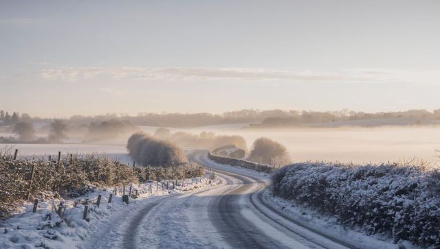 Winding snowy country road into misty valley at sunrise with frost-covered hedgerows