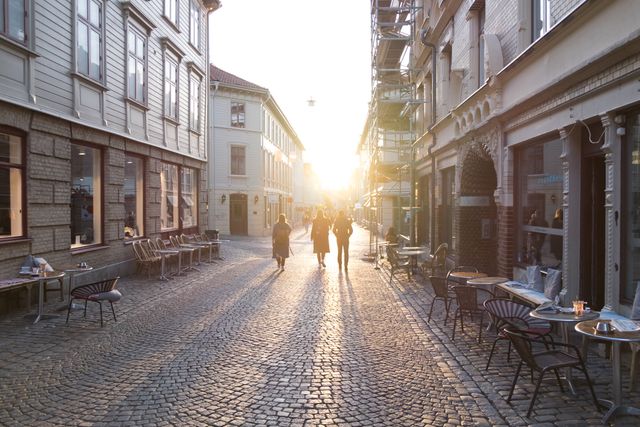 People are walking down a picturesque cobblestone street during a serene sunset in a European city. The buildings feature beautiful historic architecture with outdoor cafes and shops lining the sides of the street. This image evokes a sense of tranquility and leisure, perfect for illustrating travel destinations, urban life, and historic city experiences. Ideal for use in travel blogs, tourism advertising, city guides, and cultural websites.