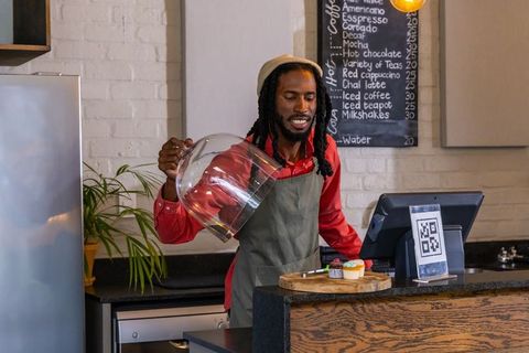 African american barista lifting cloche revealing cupcakes at cafe counter with qr pos