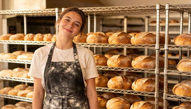 Young female baker smiling in flour-dusted apron with golden sourdough loaves on racks